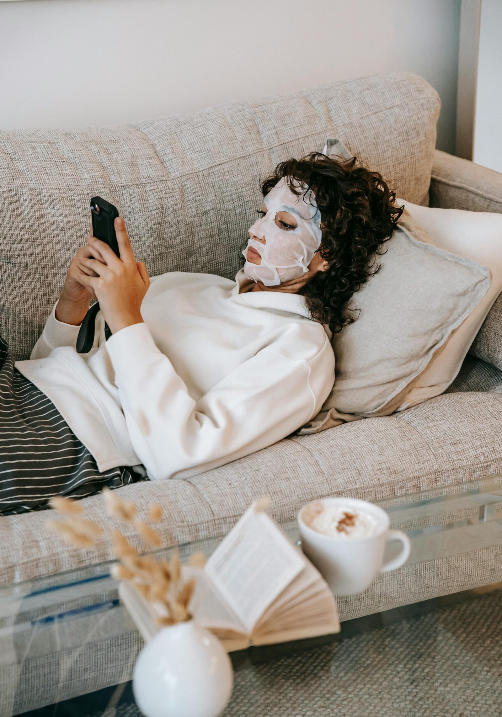 A woman relaxing on a sofa with a sheet mask, using a smartphone for leisure.