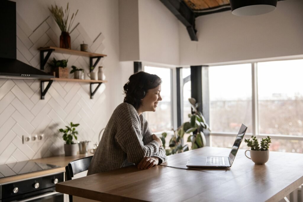 A woman enjoys a video call on her laptop in a modern, cozy kitchen setting with natural light.